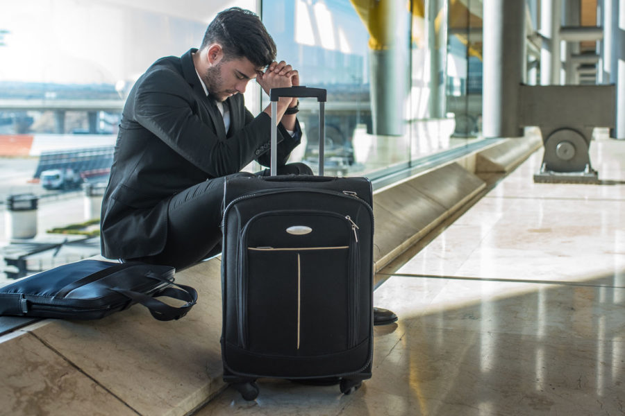 Den Jetlag bewältigen: Junger Mann im Businessanzug sitzt am Flughafen neben seinem Koffer und kann nicht schlafen