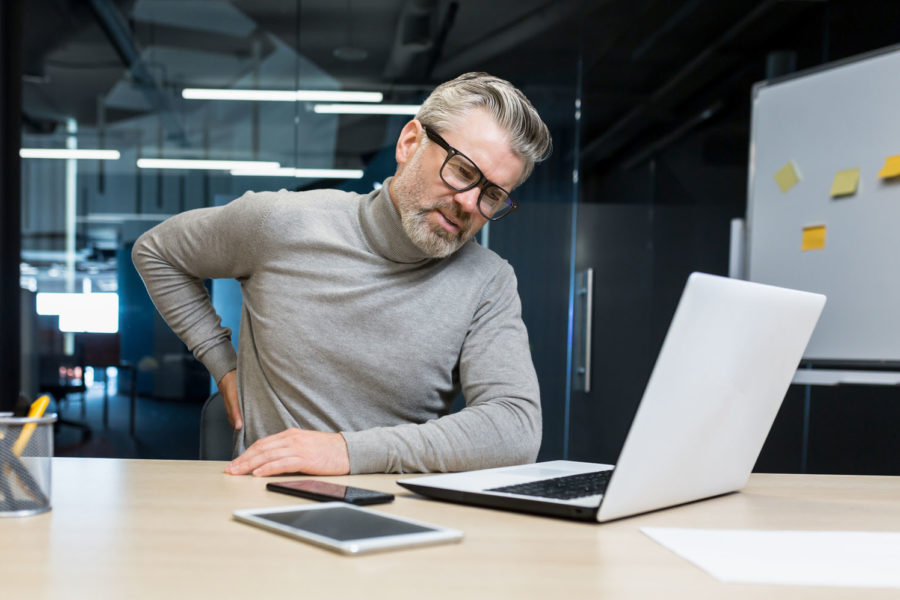 Ein älterer weißer Mann mit Brille und schmerzverzerrtem Gesicht sitzt in einem Büro vor einem Laptop und greift mit der rechten Hand in Richtung seiner Niere.