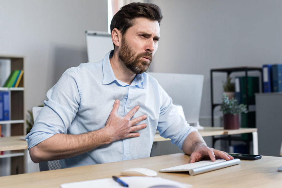 Ein Mann mittleren Alters sitzt mit schmerzverzerrtem Gesichtsausdruck an einem Schreibtisch in einer Büroumgebung. Er greift sich mit der rechten Hand an die Herzgegend.