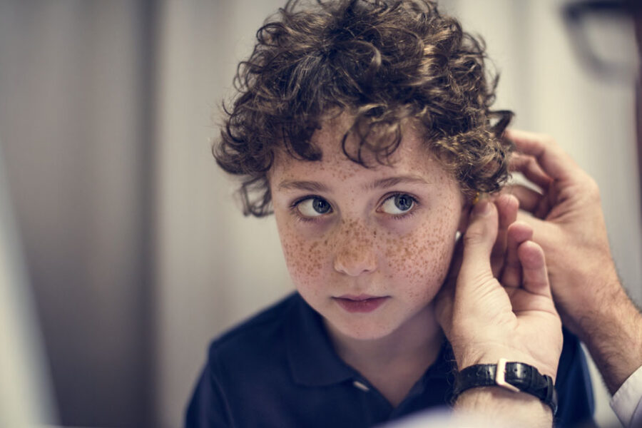 Kind mit lockigen Haaren sitzt ruhig, während eine erwachsene Person behutsam das Ohr untersucht. Die Szene zeigt eine ärztliche Ohruntersuchung bei Verdacht auf eine Mittelohretzündung (Otitis media) im medizinischen Umfeld.